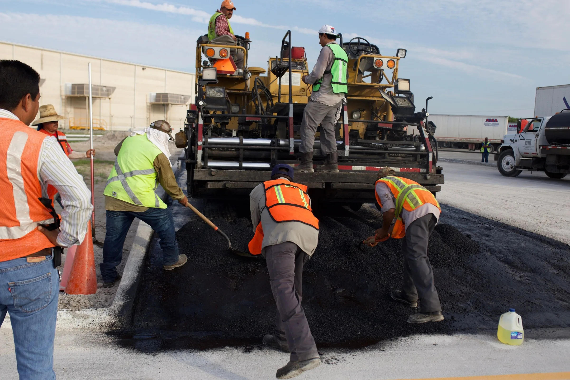 Road construction crew in safety vests spreading asphalt from a paving truck onto a parking lot.