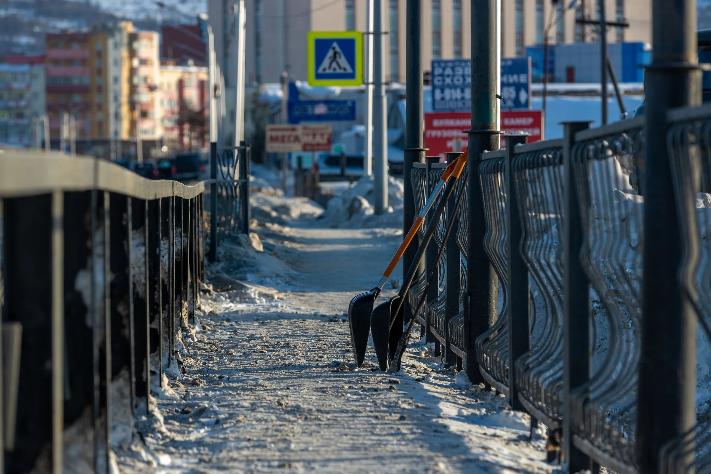 Snow removal worker with shovel clearing snow from narrow pedestrian pathway between metal railings in urban area.