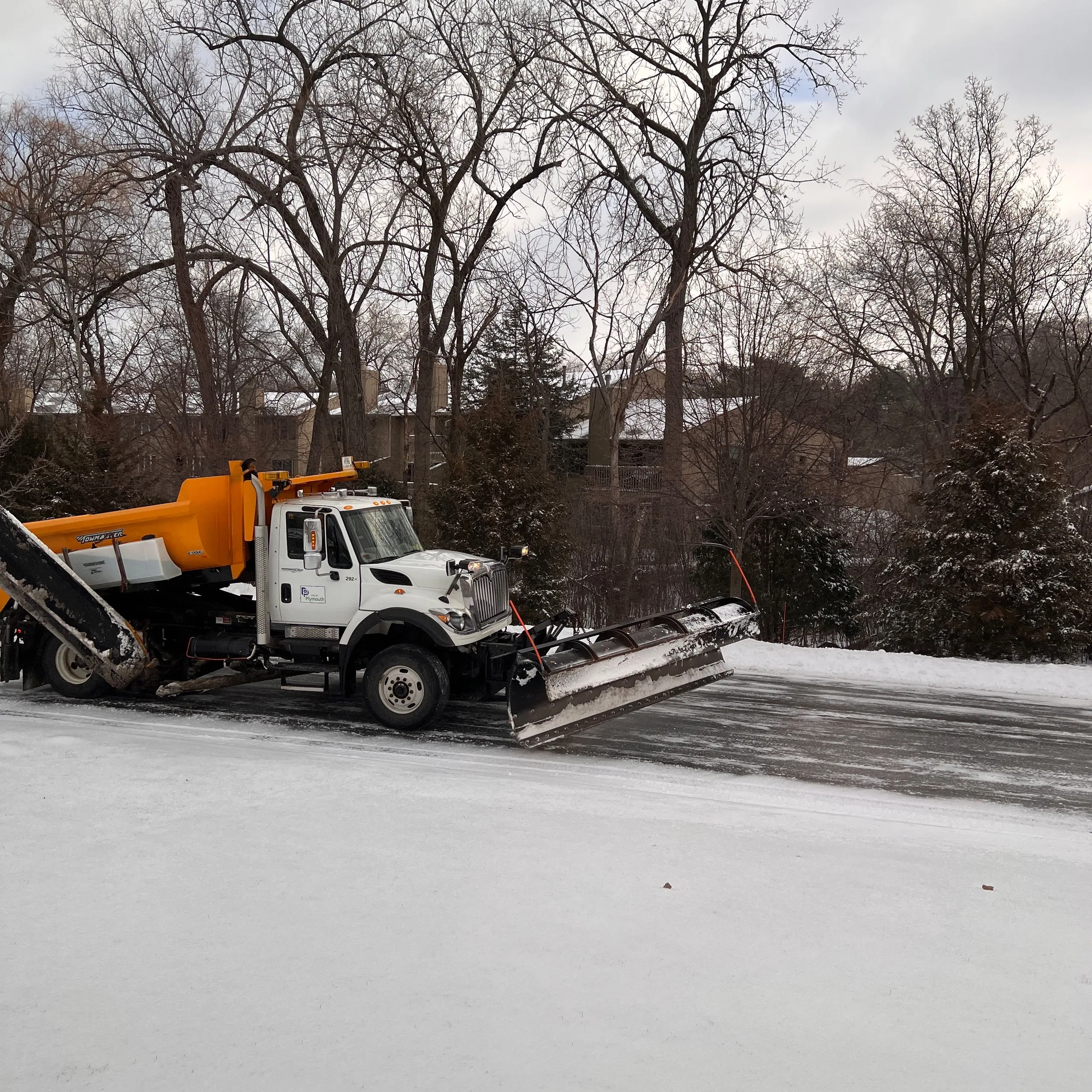 White and orange snow plow truck clearing ice from a residential driveway in winter.