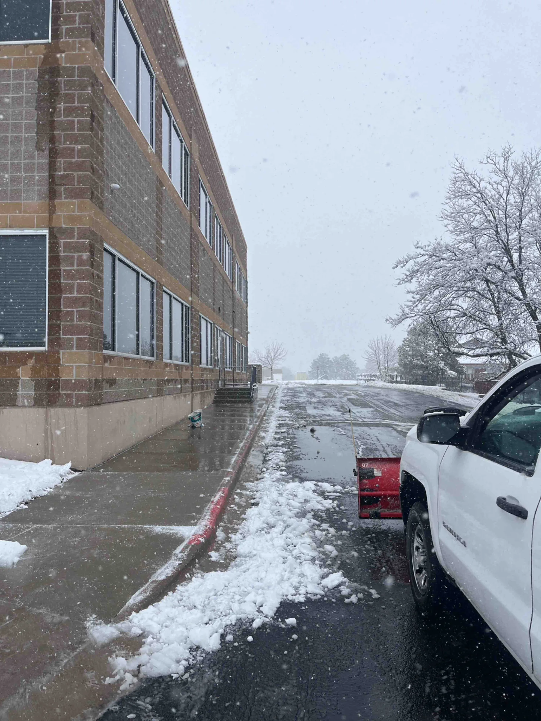 White pickup truck parked beside brick building during active snowfall in winter parking lot.