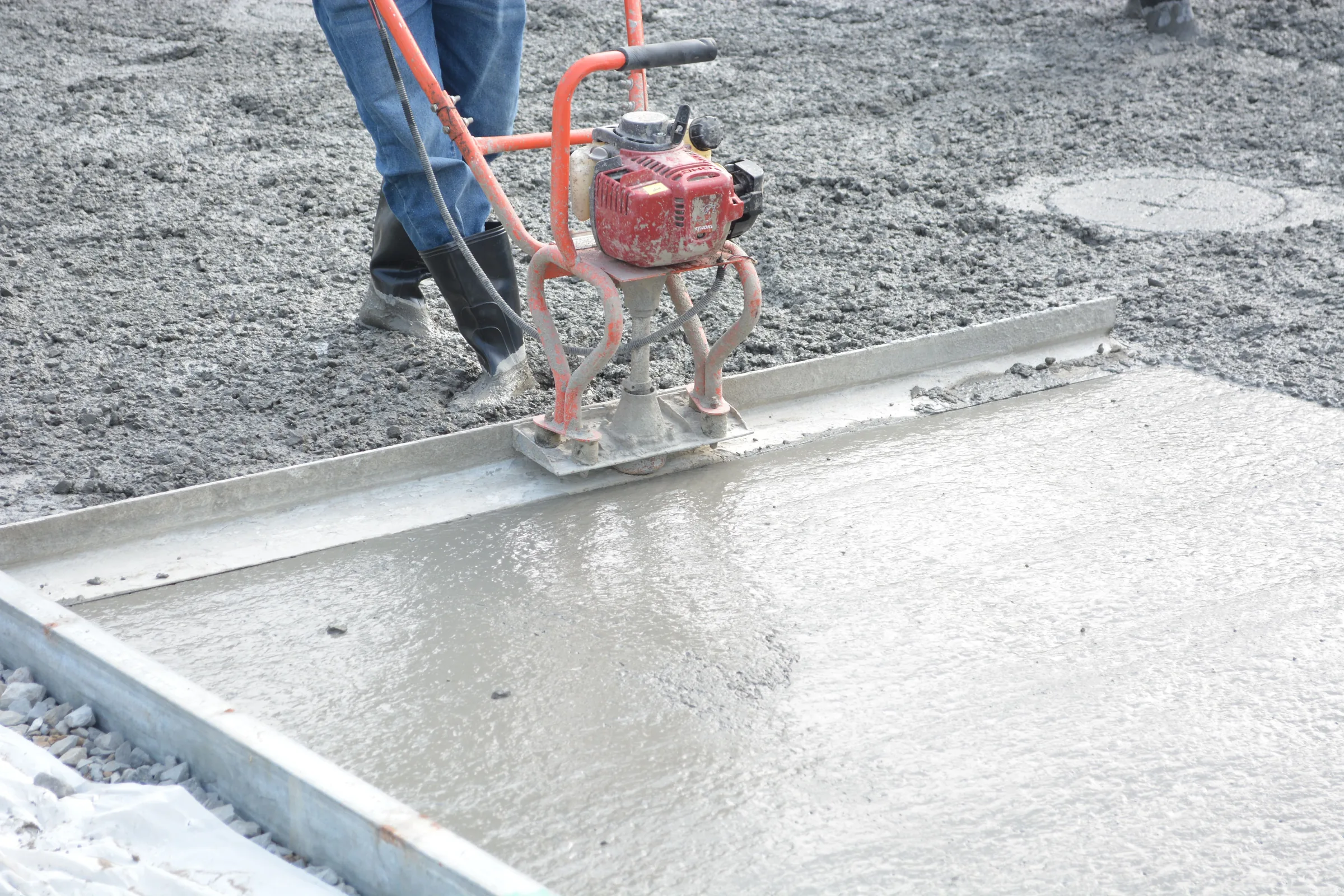 Worker operating a red concrete power screed machine on wet concrete surface during construction.