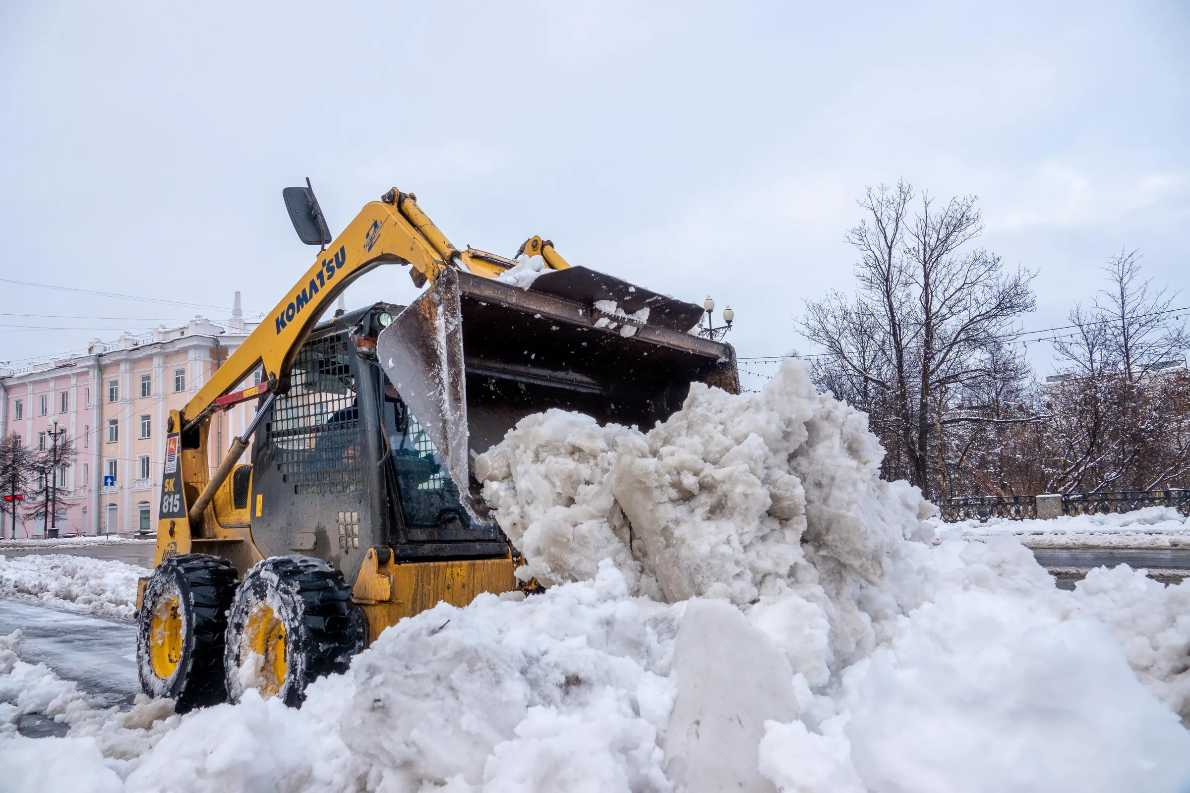 Yellow Komatsu snow removal truck dumping large pile of snow in urban area.